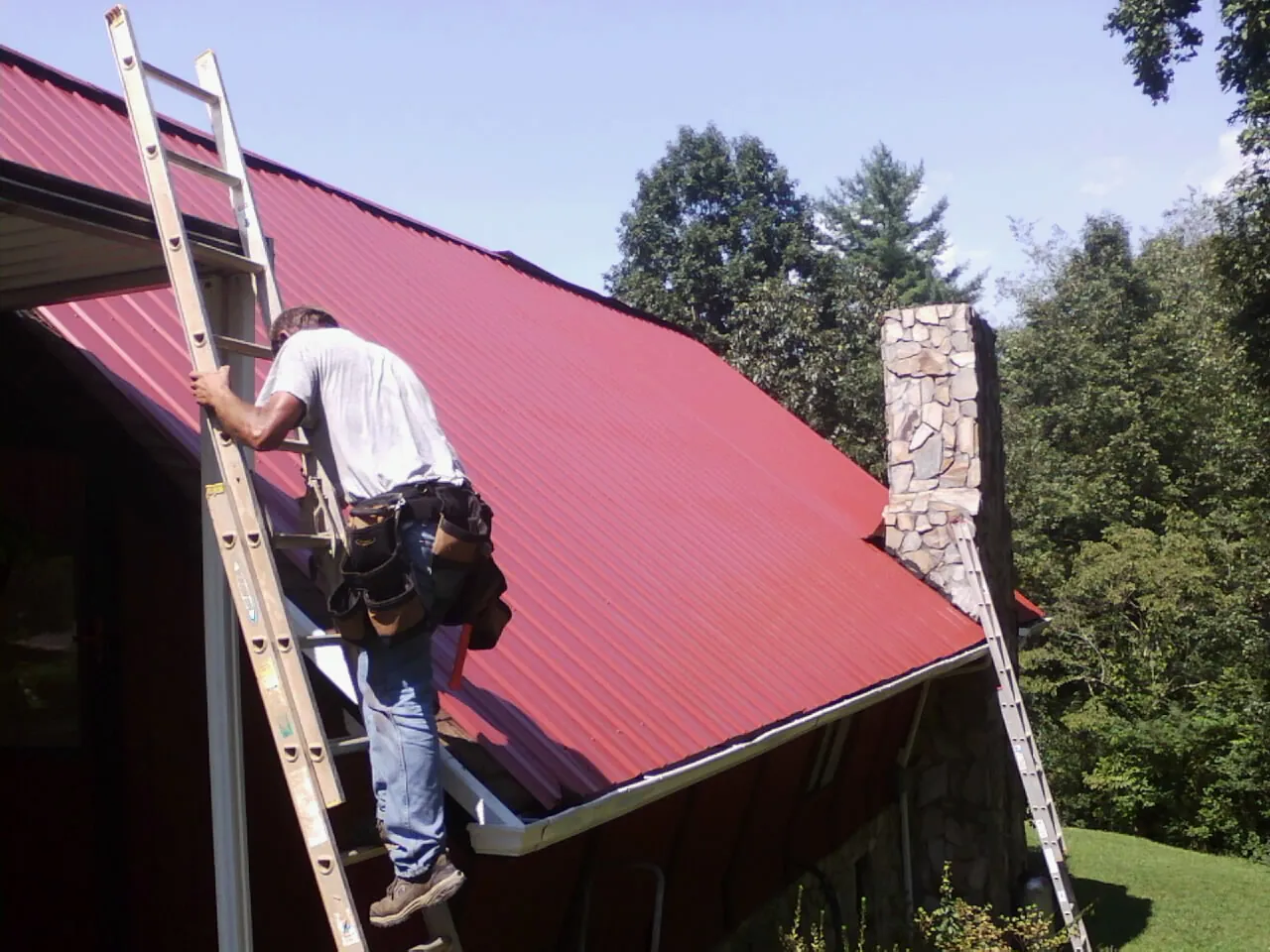 Roofer on a ladder beside a red metal roof and stone chimney — Soffit & Fascia Repair by Roofing Brothers in Port Hadlock